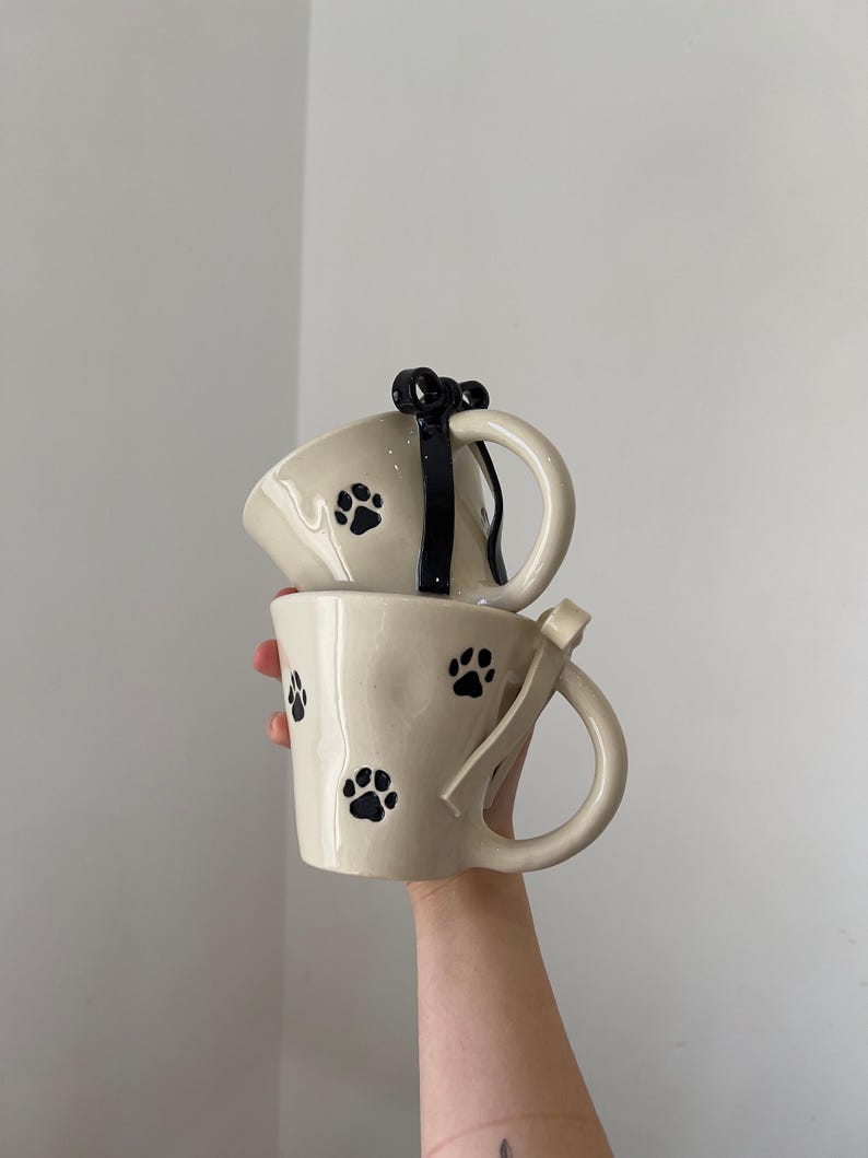 May include: Two cream-colored ceramic mugs with black paw prints. One mug has a black bow detail on the handle, and the other has a cream-colored ribbon detail. The mugs are stacked on top of each other.