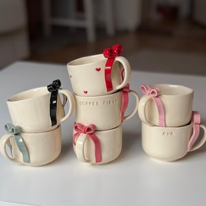 May include: A stack of cream-coloured ceramic mugs, each adorned with a colourful bow. Some mugs feature red hearts, the text "COFFEE FIRST", or the word "MUM". Bows are in red, black, pink, and teal.