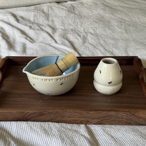 May include: A ceramic matcha bowl with a light blue interior and cream exterior featuring small blue floral accents and the embossed text "BUT FIRST, MATCHA". A bamboo whisk rests inside the bowl, accompanied by a matching cream ceramic whisk holder.
