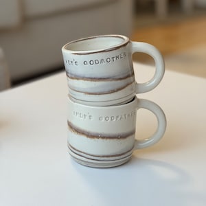 May include: Two cream-glazed ceramic mugs stacked on top of each other, featuring brown stripe detailing. The top mug is inscribed with "INDY'S GODMOTHER," and the bottom mug reads "INDY'S GODFATHER." Each mug has a rounded handle.