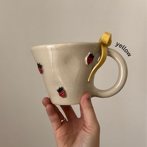 May include: Cream-colored ceramic mug with a curved handle and a yellow accent. The mug is adorned with small, red strawberry designs. The word "yellow" is written on the side of the mug.
