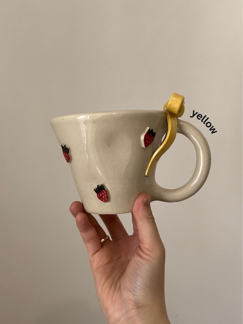 May include: Cream-colored ceramic mug with a curved handle and a yellow accent. The mug is adorned with small, red strawberry designs. The word "yellow" is written on the side of the mug.