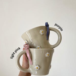 May include: Two beige ceramic mugs with daisy embellishments and large handles. One mug has a light pink ribbon, the other a purple ribbon. The words "light pink" and "purple" are written on the image.