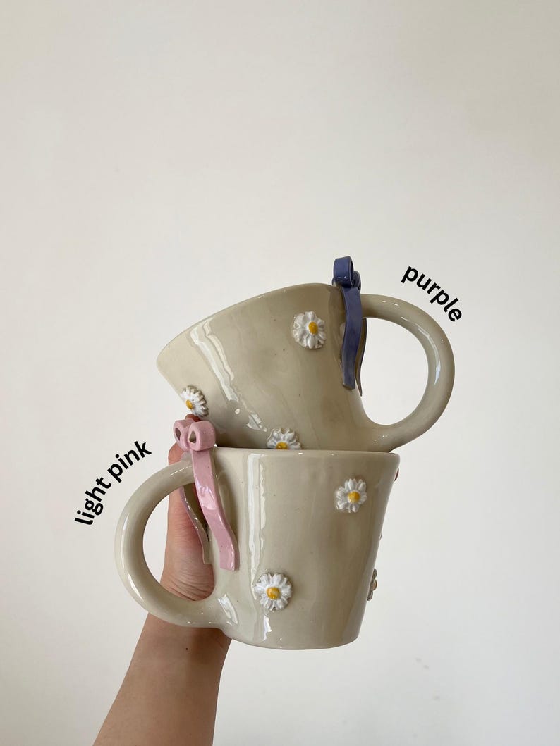 May include: Two beige ceramic mugs with daisy embellishments and large handles. One mug has a light pink ribbon, the other a purple ribbon. The words "light pink" and "purple" are written on the image.