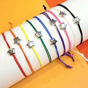 May include: A set of colorful string bracelets with silver star charms. The bracelets are arranged in a rainbow order, with red, orange, yellow, green, blue, purple, pink, white, and black.