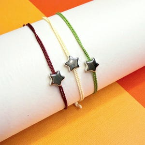 Puede incluir: Tres pulseras de cuerda con dijes de estrella plateada. Las pulseras son de color burdeos, crema y verde.