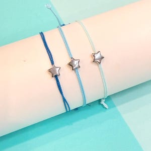 May include: Three blue friendship bracelets with silver star charms. The bracelets are tied on a white surface with a blue background.