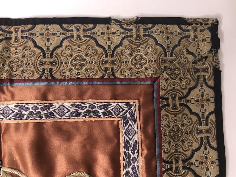 Puede incluir: Un primer plano de un tapiz con fondo marr&oacute;n y un borde multicolor. El borde presenta un patr&oacute;n geom&eacute;trico con acentos azules, rojos y dorados. El tapiz est&aacute; hecho de una tela tejida y tiene una superficie texturizada.