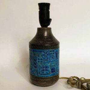 Può includere: Una lampada da tavolo in ceramica con base marrone scuro e sezione a motivi blu turchese. La lampada ha un portalampada nero e un cavo color oro. La sezione turchese presenta un motivo geometrico ripetuto.
