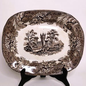 May include: A brown and white oval ceramic serving platter with a detailed floral border and a central image of a black and white castle in a landscape.