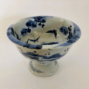 May include: A small, white ceramic bowl with a blue and white floral design. The bowl has a pedestal base and is decorated with a floral pattern and birds.