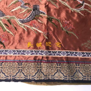 Puede incluir: Un tapiz de seda marr&oacute;n con una imagen bordada de la Gran Muralla China. El tapiz tiene un borde dorado con el texto "长城 纪念" en caracteres chinos.