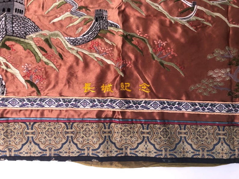 Puede incluir: Un tapiz de seda marr&oacute;n con una imagen bordada de la Gran Muralla China. El tapiz tiene un borde dorado con el texto "长城 纪念" en caracteres chinos.