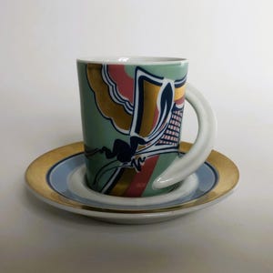 May include: A decorative ceramic mug and saucer set. The mug features a curved handle and a design with gold, pink, and blue accents on a light green background. The saucer has a gold rim and a blue center.