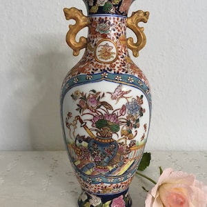 May include: A white porcelain vase with gold handles and a colorful floral design. The vase has a blue and white base with a red and gold rim.