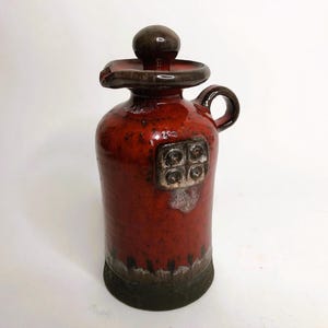 Puede incluir: Una jarra de cerámica roja con tapa marrón y un diseño decorativo. La jarra tiene un asa y un pico.