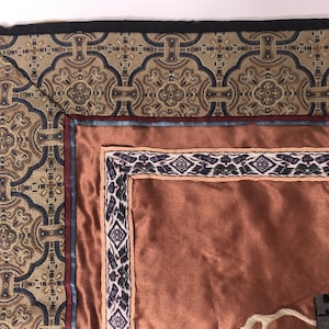 Puede incluir: Primer plano de la esquina de un tapiz con un borde estampado en marr&oacute;n, dorado y azul. El tapiz tiene un respaldo de sat&eacute;n marr&oacute;n y un borde m&aacute;s peque&ntilde;o e intrincado en tonos de morado, blanco y negro.