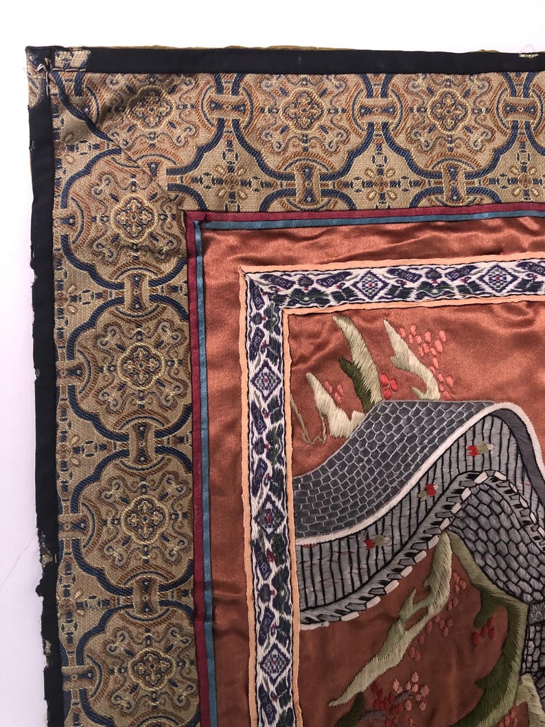 Puede incluir: Primer plano de una esquina de un tapiz con un dise&ntilde;o bordado detallado. El tapiz presenta un fondo marr&oacute;n con un puente gris y &aacute;rboles verdes. El borde es una tela estampada en oro y azul.