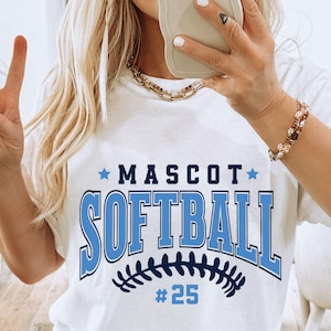 Könnte beinhalten: Weißes T-Shirt mit den Worten "MASCOT SOFTBALL" in blauen Blockbuchstaben. Die Nummer 25 steht unter dem Wort Softball. Das Shirt hat eine Baseball-Grafik am unteren Rand des Wortes Softball.