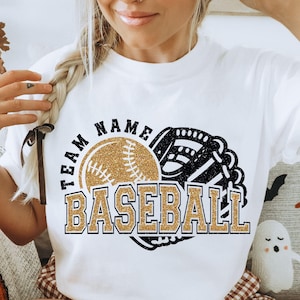 Może przedstawiać: Biała koszulka z grafiką baseballową. Projekt przedstawia złotą brokatową piłkę i rękawicę do baseballu, ze słowem "BASEBALL" w złotych brokatowych literach. "TEAM NAME" jest wygięte nad piłką.