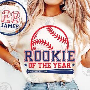 May include: A cream-coloured t-shirt featuring a red baseball graphic, the text ROOKIE OF THE YEAR in blue, and a blue baseball bat. An inset shows the back of the shirt with the number 28 in red baseball-stitch font and the name JAMES in blue.
