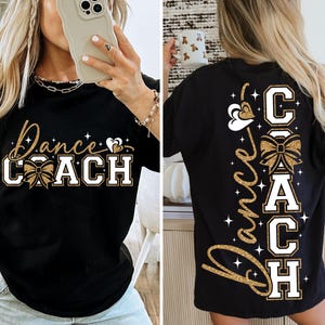 May include: Black t-shirt with the words "Dance Coach" in gold glitter script and block letters. The front design includes a bow and heart accents. The back of the shirt repeats the text vertically with a bow and star accents.