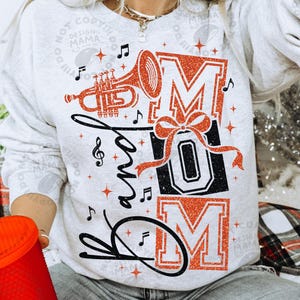 May include: A light grey crewneck sweatshirt featuring a graphic print with the text "band MOM". The design includes a glittery orange trumpet, a glittery orange bow, and black musical notes, with the word "band" in black script.