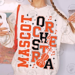 May include: A cream-coloured crewneck sweatshirt featuring the orange and black text MASCOT ORCHESTRA. The design includes musical notes and a violin graphic, accented by black star shapes. The sweatshirt has long sleeves and a ribbed neckline.