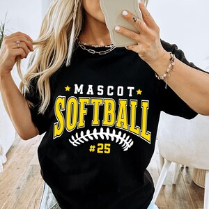 May include: A black short-sleeved t-shirt featuring the yellow text "MASCOT" above the word "SOFTBALL" in a large collegiate font. Below the text is a white graphic of a softball stitch pattern and the yellow number "#25" centered.