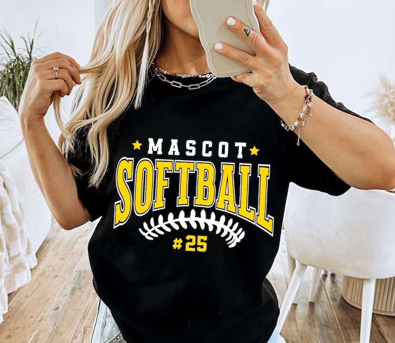 May include: A black short-sleeved t-shirt featuring the yellow text "MASCOT" above the word "SOFTBALL" in a large collegiate font. Below the text is a white graphic of a softball stitch pattern and the yellow number "#25" centered.