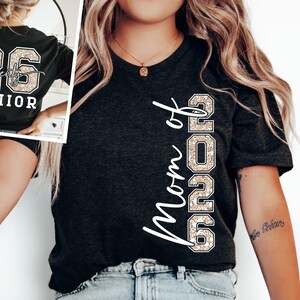 May include: Dark gray t-shirt with the text "Mom of 2026" in white script. The number "2026" is in a glittery, tan font. The back of the shirt in the inset image says "26 Twenty SENIOR" in a similar glittery font.