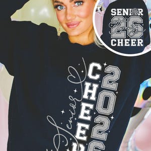 May include: Black sweatshirt with the words "Senior Cheer 2026" in white and silver glitter lettering. The back of the sweatshirt has the words "Senior Twenty 26 Cheer" with a cheerleader graphic. The sweatshirt is worn by a person.