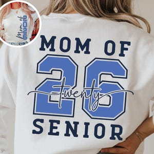 May include: White sweatshirt with the text "MOM OF 26 SENIOR" in navy blue and light blue. The number "26" is in a bold, outlined font. The word "twenty" is written in cursive between the numbers.