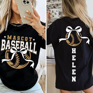 Peut inclure: T-shirt de baseball noir avec un motif dor&eacute; et blanc. Le devant affiche "MASCOT BASEBALL" au-dessus d'un graphisme de baseball avec un n&oelig;ud blanc. Le dos pr&eacute;sente un graphisme de baseball avec un n&oelig;ud et le nom "HELEN".