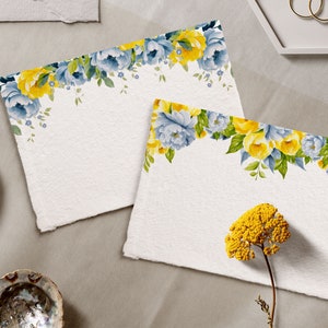 May include: Two blank white cards with a floral border of blue and yellow flowers. The cards are on a light gray surface with other decorative items.