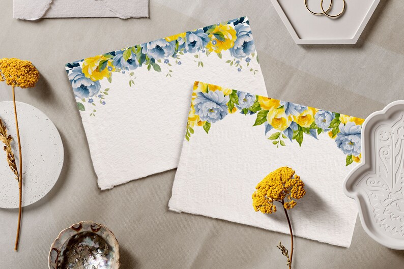 May include: Two blank white cards with a floral border of blue and yellow flowers. The cards are on a light gray surface with other decorative items.