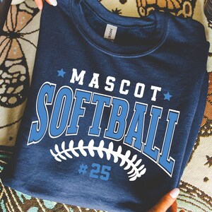 May include: A navy blue crewneck sweatshirt featuring the white and light blue text MASCOT SOFTBALL with two small stars. Below the text is a white graphic of softball stitching and the number #25 in light blue. The garment has a Gildan label.