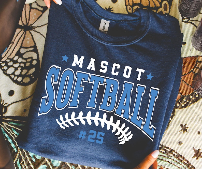 May include: A navy blue crewneck sweatshirt featuring the white and light blue text MASCOT SOFTBALL with two small stars. Below the text is a white graphic of softball stitching and the number #25 in light blue. The garment has a Gildan label.