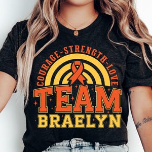 May include: A dark charcoal grey t-shirt featuring a graphic design with a yellow rainbow, an orange awareness ribbon, and the text COURAGE-STRENGTH-LOVE TEAM BRAELYN in orange and yellow block lettering with small star accents throughout the print.
