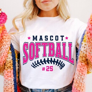 May include: A white short-sleeve t-shirt featuring the text MASCOT SOFTBALL in dark blue and pink, with two pink stars, a dark blue softball stitch graphic, and the number 25 in pink below the text, worn under a chunky knit cardigan.
