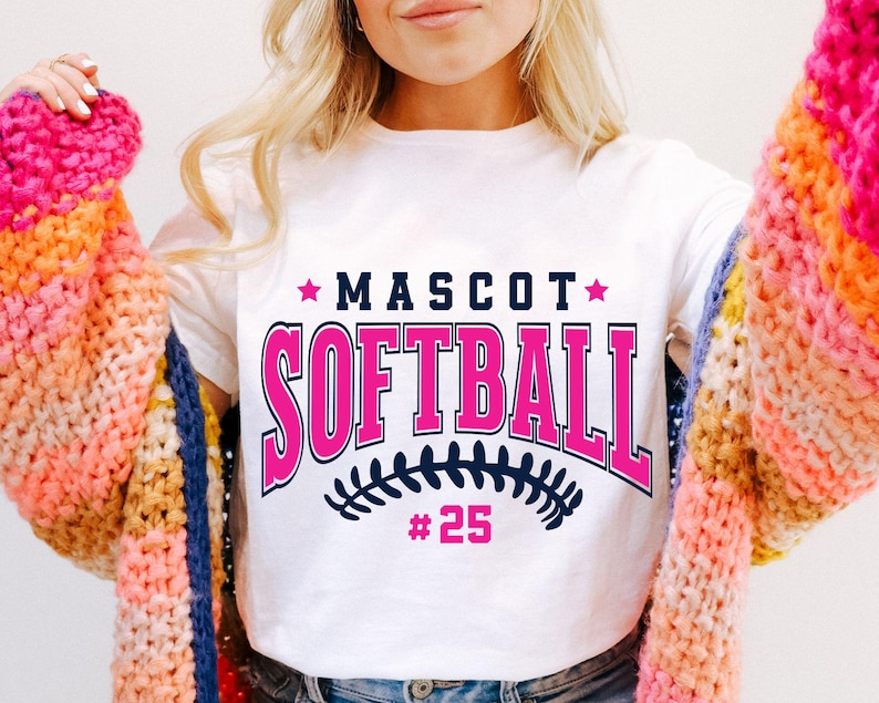 May include: A white short-sleeve t-shirt featuring the text MASCOT SOFTBALL in dark blue and pink, with two pink stars, a dark blue softball stitch graphic, and the number 25 in pink below the text, worn under a chunky knit cardigan.