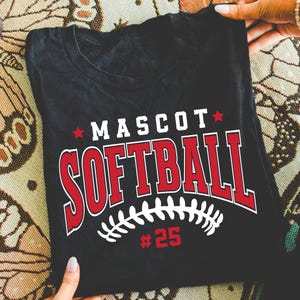 May include: A black t-shirt featuring a graphic print with the word MASCOT in white letters, the word SOFTBALL in large red letters with white outlines, a white softball stitch design, and the number 25 in red, accented by two small red stars.