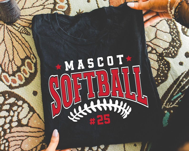 May include: A black t-shirt featuring a graphic print with the word MASCOT in white letters, the word SOFTBALL in large red letters with white outlines, a white softball stitch design, and the number 25 in red, accented by two small red stars.