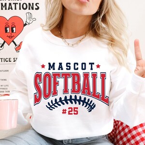 May include: A white crewneck sweatshirt featuring the text MASCOT SOFTBALL in navy blue and red collegiate lettering. Below the text is a navy blue graphic of softball stitching and the red number 25. The design is centered on the chest.