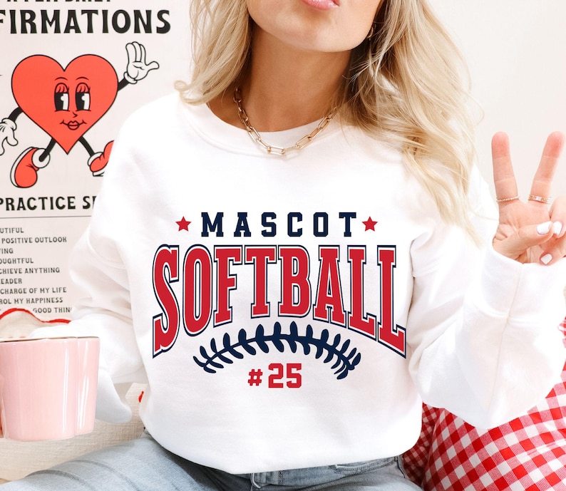 May include: A white crewneck sweatshirt featuring the text MASCOT SOFTBALL in navy blue and red collegiate lettering. Below the text is a navy blue graphic of softball stitching and the red number 25. The design is centered on the chest.