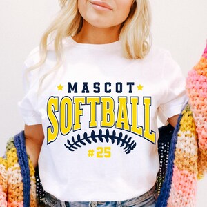 May include: A white short-sleeved t-shirt featuring the text MASCOT SOFTBALL in dark blue and yellow block lettering, with a dark blue softball stitch graphic and the number 25 in yellow below the text, accented by two small yellow stars.