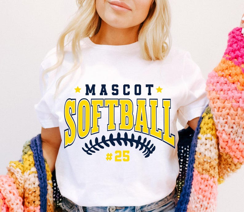 May include: A white short-sleeved t-shirt featuring the text MASCOT SOFTBALL in dark blue and yellow block lettering, with a dark blue softball stitch graphic and the number 25 in yellow below the text, accented by two small yellow stars.