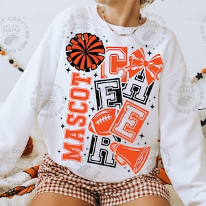 May include: A white long-sleeve sweatshirt featuring a graphic print in orange and black. The design includes the word MASCOT vertically, the word CHEER in block letters, a pom-pom, a hair bow, a football, and a megaphone with star accents.