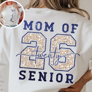 May include: White sweatshirt with the text "MOM OF 26 SENIOR" in navy blue. The number "26" is filled with a glittery gold material. The word "twenty" is written in cursive font.