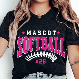 May include: A dark charcoal gray short-sleeve t-shirt featuring the text MASCOT SOFTBALL in bold pink letters with white outlines. Below the text is a white graphic of softball stitching and the pink text #25, flanked by two pink stars.